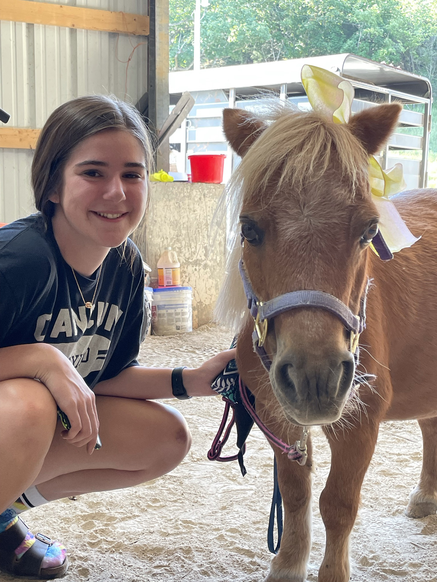 A smiling girl kneels beside a small brown pony with a light mane and a yellow and white ribbon on its head, inside a barn with sunlight streaming in from outside.