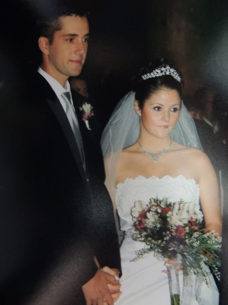 Emeri and JR stand side by side, holding hands. Emeri wears a white strapless gown, tiara, veil, and holds a bouquet; JR wears a dark suit with a boutonnière. Both appear solemn.