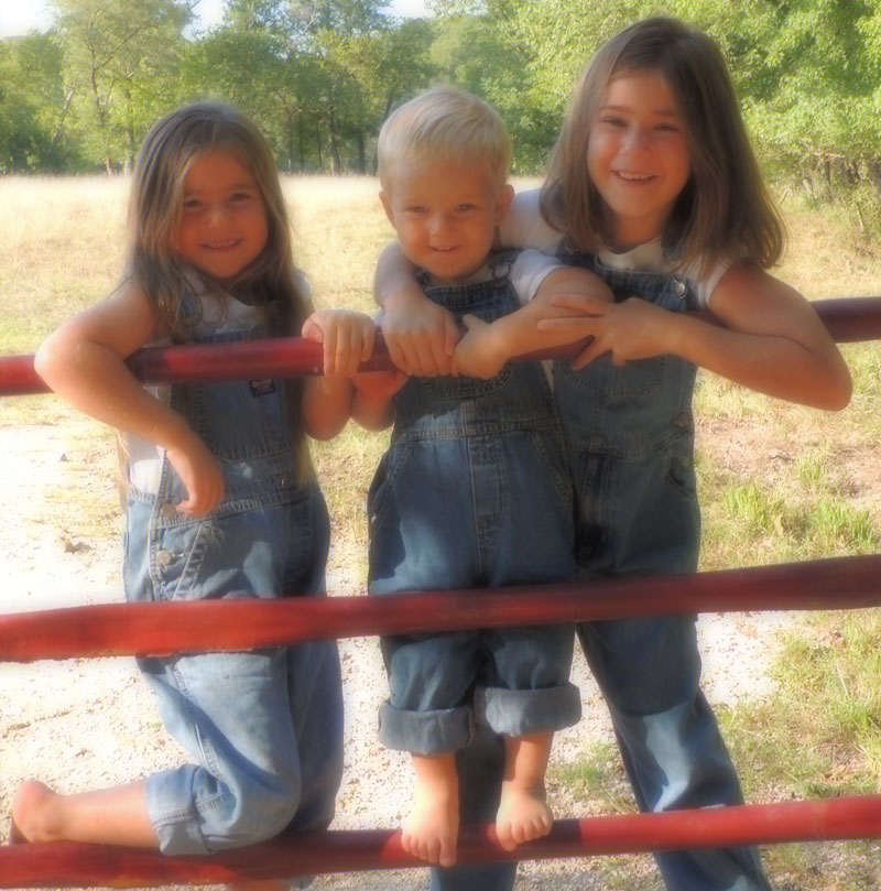 Three young children wearing denim overalls smile and pose together on a red metal fence outdoors in a sunlit, grassy area with trees in the background.