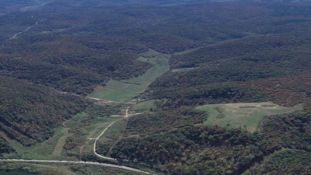 Aerial view of rolling, forested hills with green clearings and winding dirt roads cutting through the landscape. The area is lush with trees, and some open grassy patches are visible among the woods.