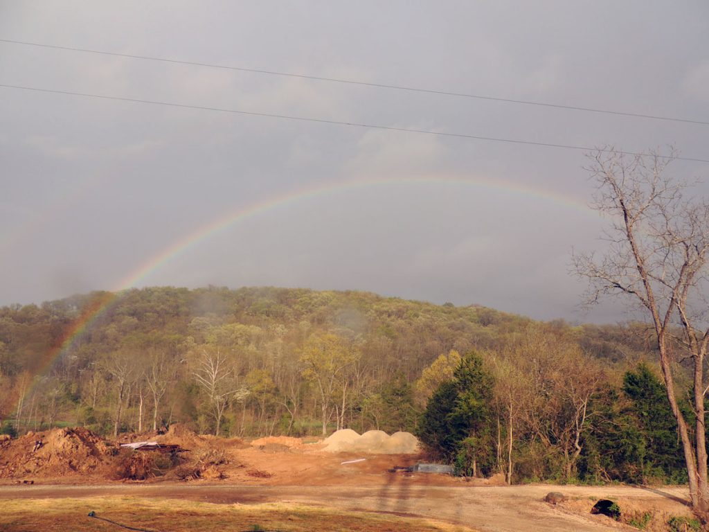 A faint rainbow arcs over a forested hillside under a cloudy sky, with bare and green trees. Below, there is a cleared, brown dirt area with piles of soil, possibly part of a construction or landscaping site.