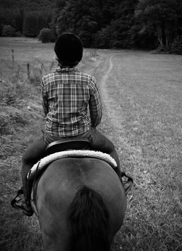A person wearing a plaid shirt and helmet rides a horse along a grassy path bordered by trees, viewed from behind in black and white.