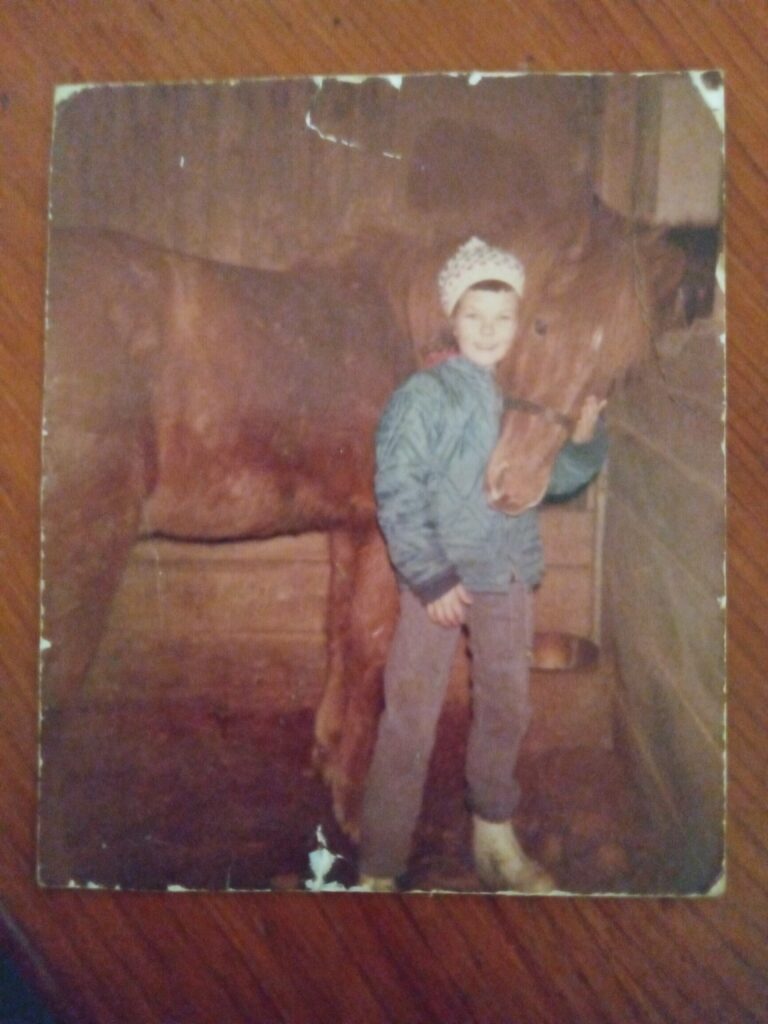 A young child in a blue jacket and jeans stands smiling beside a brown horse in a wooden stall, gently holding the horse’s head. The photo has a vintage look with some wear on its edges.